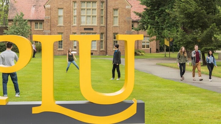 A large yellow PLU sign on campus, with students walking down the sidewalk beside it and other students playing frisbee on the grass behind the sign
