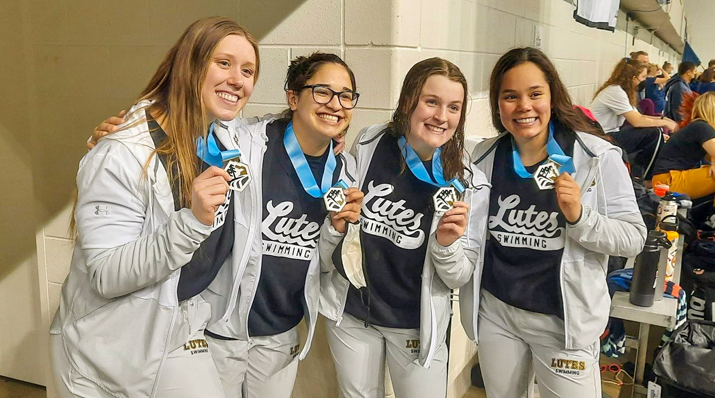 Baily Hamilton (far left) stands alongside three women swimming teammates. The four women are smiling at the camera and holding up their medals.