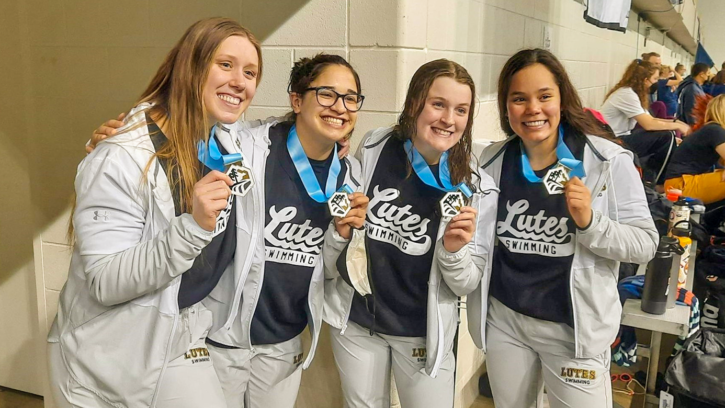 Baily Hamilton (far left) stands alongside three women swimming teammates. The four women are smiling at the camera and holding up their medals.