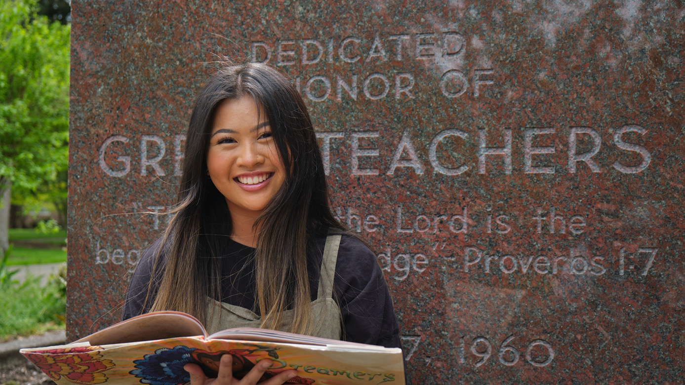 Sophia Barro smiles while holding a book in front of a monument dedicated to teachers on the PLU campus.