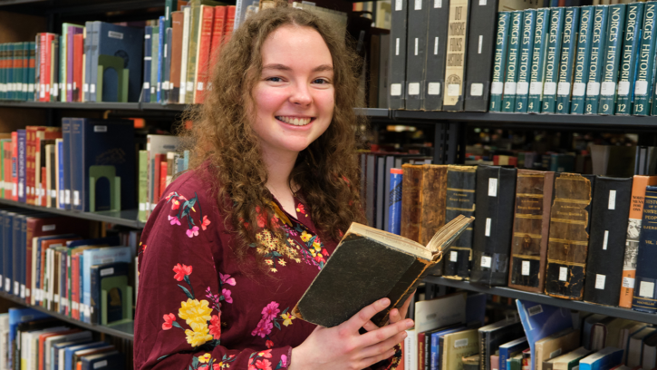 Kathryn Einan stands in front of a bookcase filled with books. She is holding a book open and is smiling at the camera.