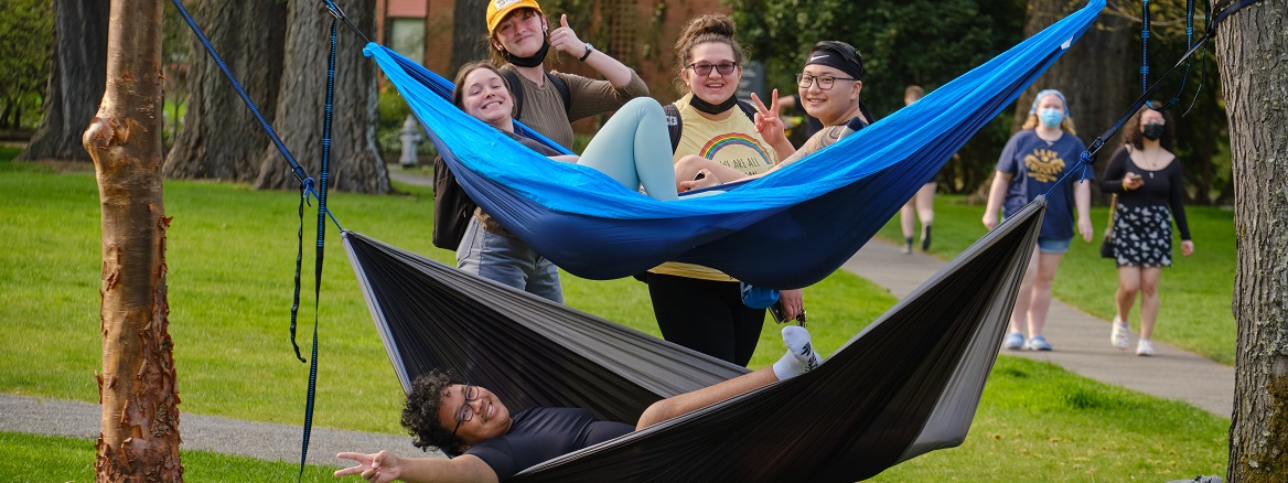 A group of students, laying in or standing by hammocks outside on campus, pose & smile for the camera