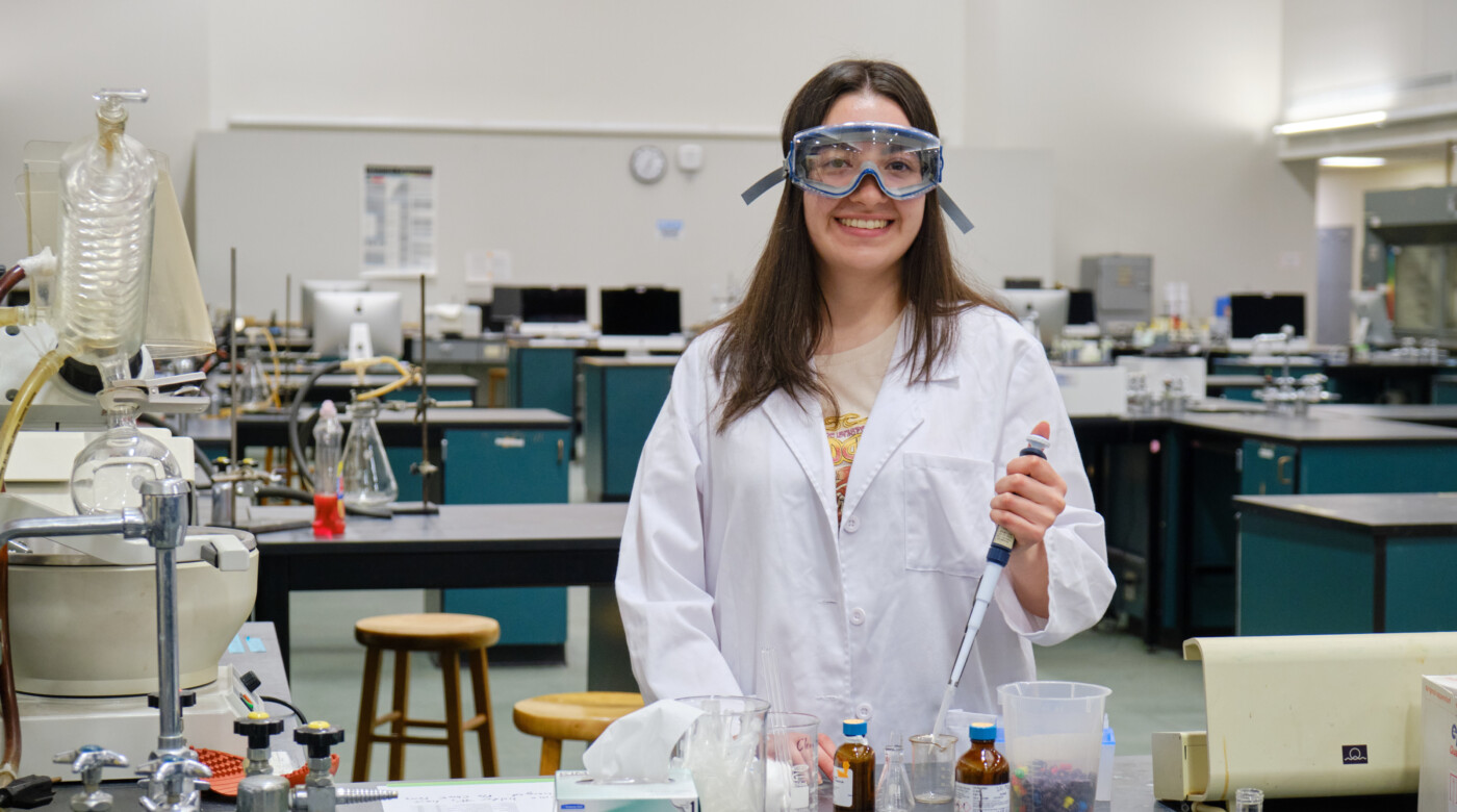 Yaquelin Rarmirez stands in a lab holding dropper over a jar. She is wearing a white lab coat and safety goggles.