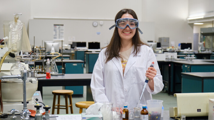 Yaquelin Rarmirez stands in a lab holding dropper over a jar. She is wearing a white lab coat and safety goggles.