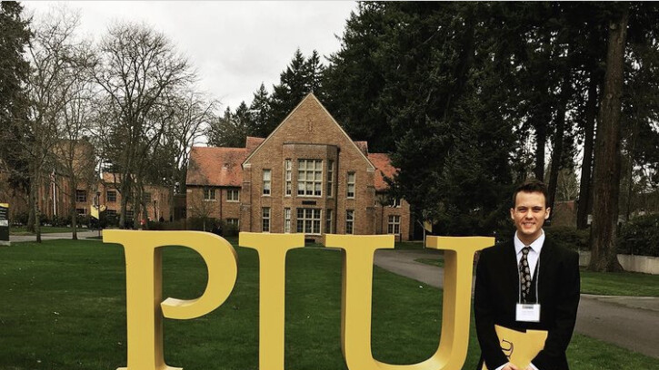 Parker Simpson stands in front of a yellow PLU sign on the university campus. He is smiling at the camera in a dark suit.