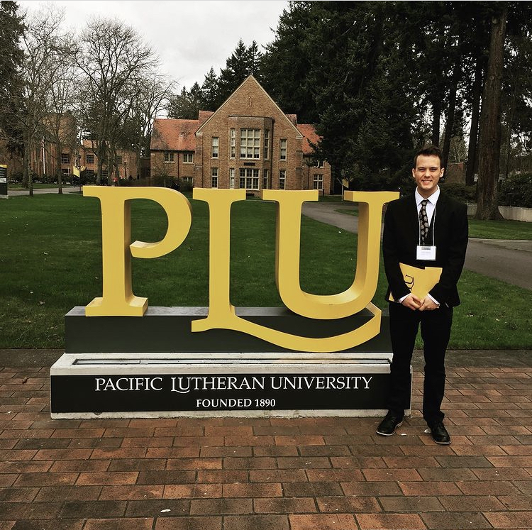 Parker Simpson stands in front of a yellow PLU sign on the university campus. He is smiling at the camera in a dark suit.