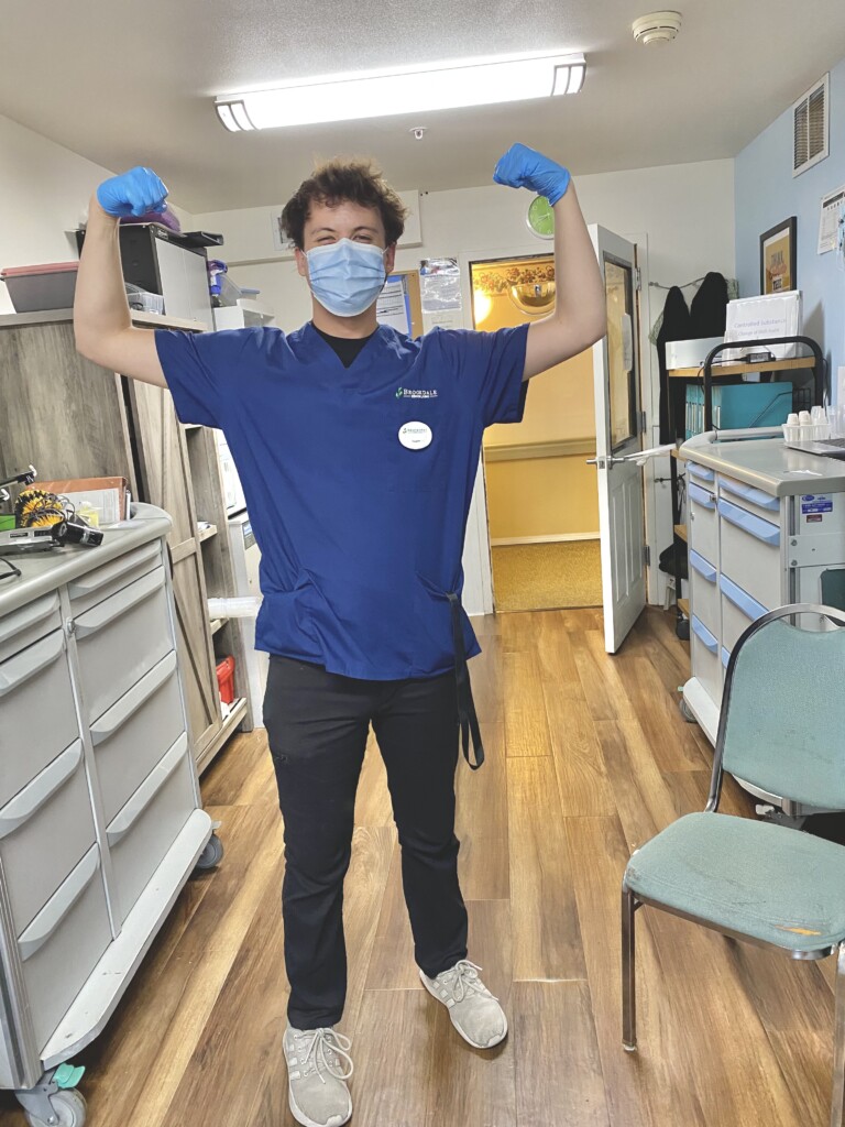 Parker Simpson stands with his hands raised while wearing his scrubs, mask and gloves in an medical office.