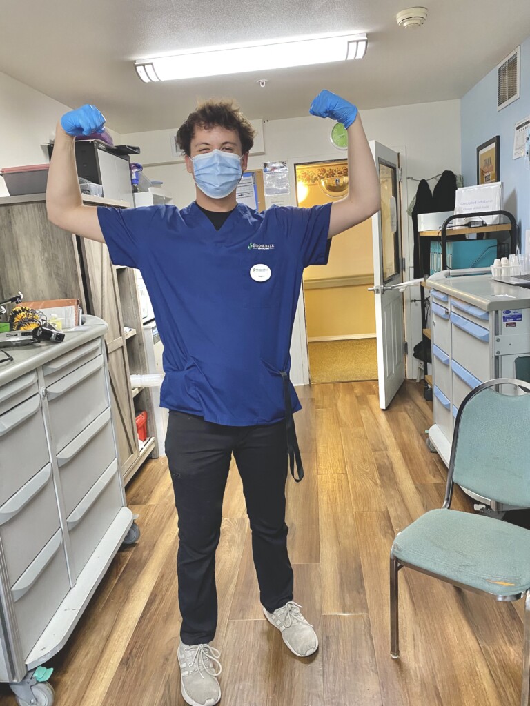 B3E116DC-96A1-44CF-938C-D65D82B8D116 Parker Simpson stands with his hands raised while wearing his scrubs, mask and gloves in an medical office.
