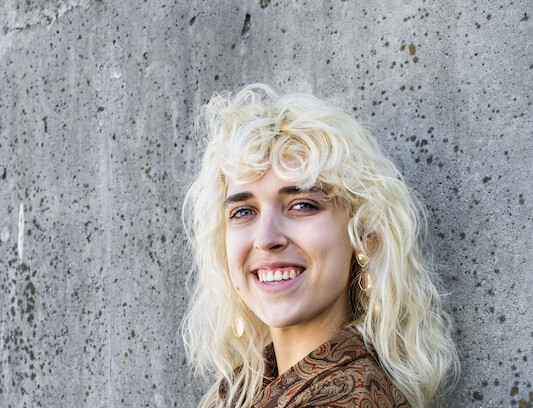 Peyton Noreen smiles at the camera while leaning against a wall. They have blonde hair and are wearing a brown shirt and pants.