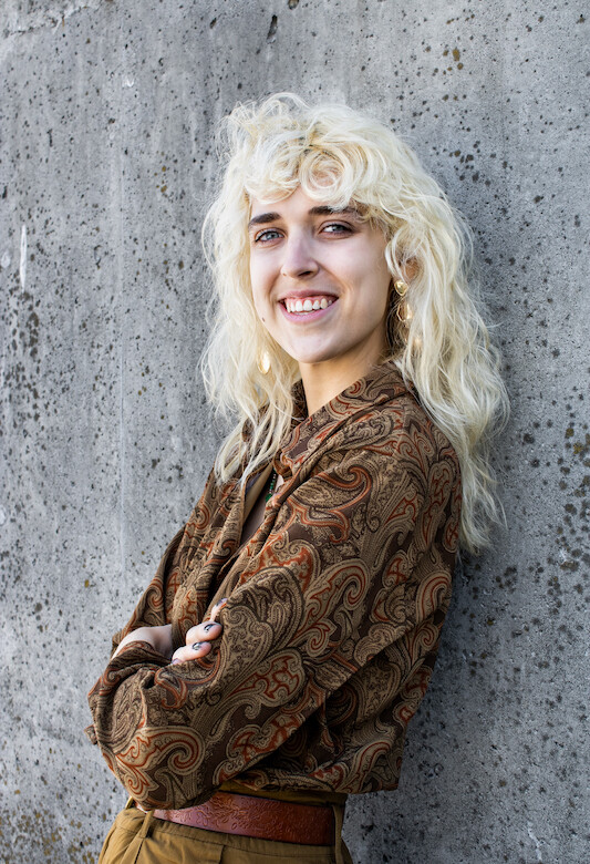 Peyton Noreen smiles at the camera while leaning against a wall. They have blonde hair and are wearing a brown shirt and pants.