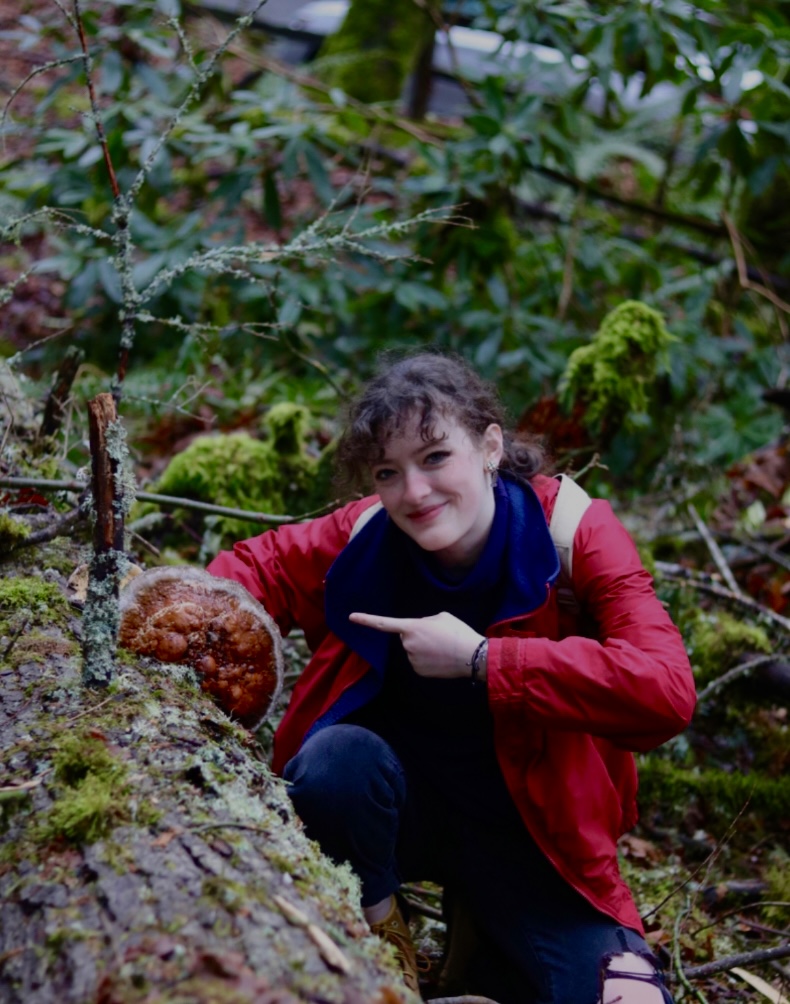 Fiona Ashton-Knochel points to a mushroom on a log in a densely green forest.