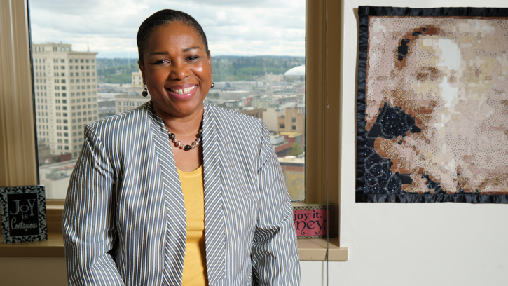 Lisa Woods smiles at the camera in her office. The window behind her looks over the city of Tacoma and there is a picture of Martin Luther King Jr. to her right.