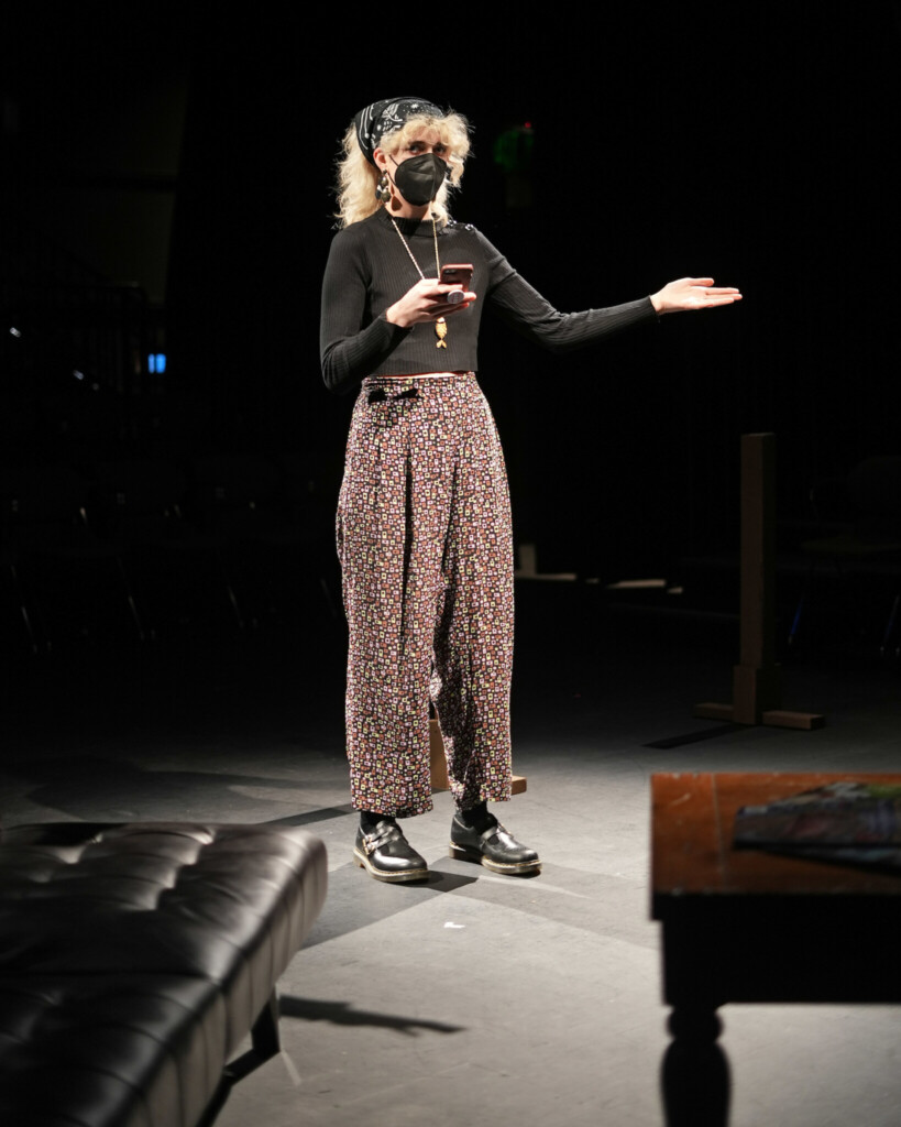 Peyton is seen on a stage wearing a mask, holding a phone and gesturing.