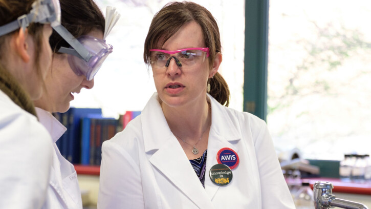 Amy Siegesmund is seen speaking to two students in a lab space. She's wearing a white coat and goggles.