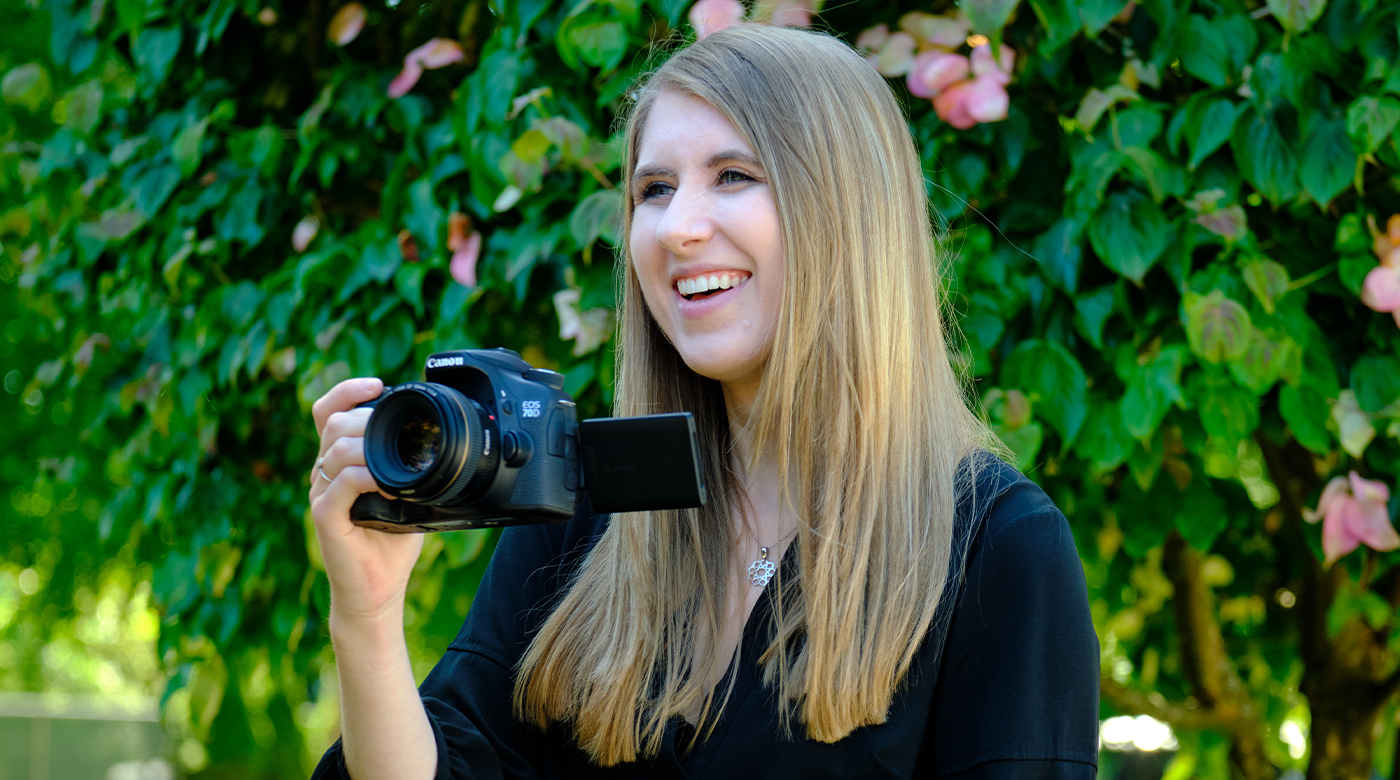 Kate Hall looks off camera while holding a camera in her right hand. She is standing in front of a flower bush, wearing a black blouse.