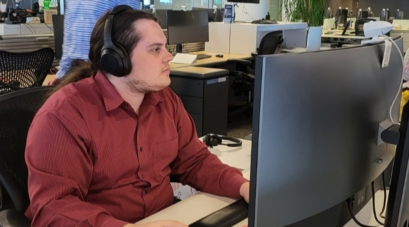 Travis McDaneld looks off to the side as he sits at a desk in front of a computer screen. He is wearing a red button up long-sleeve dress shirt and headphones.