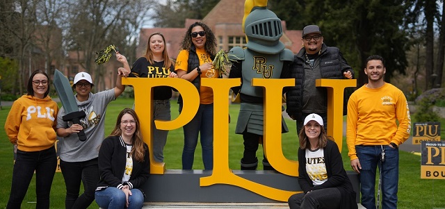 A group of 8 PLU admission counselors in Lute gear stand on and around a big yellow PLU monument sign along with the PLU mascot Lancelute