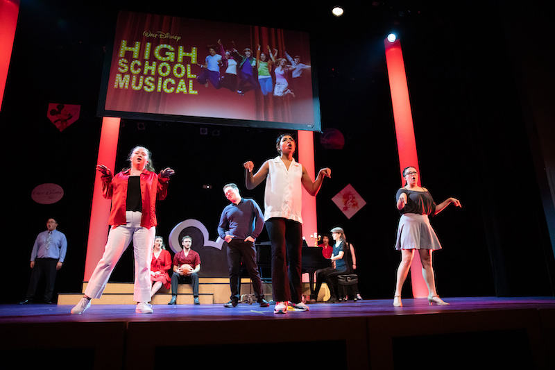 Eight actors are on a stage in various dance poses. Behind them is a giant screen with the "High School Musical" logo on it.