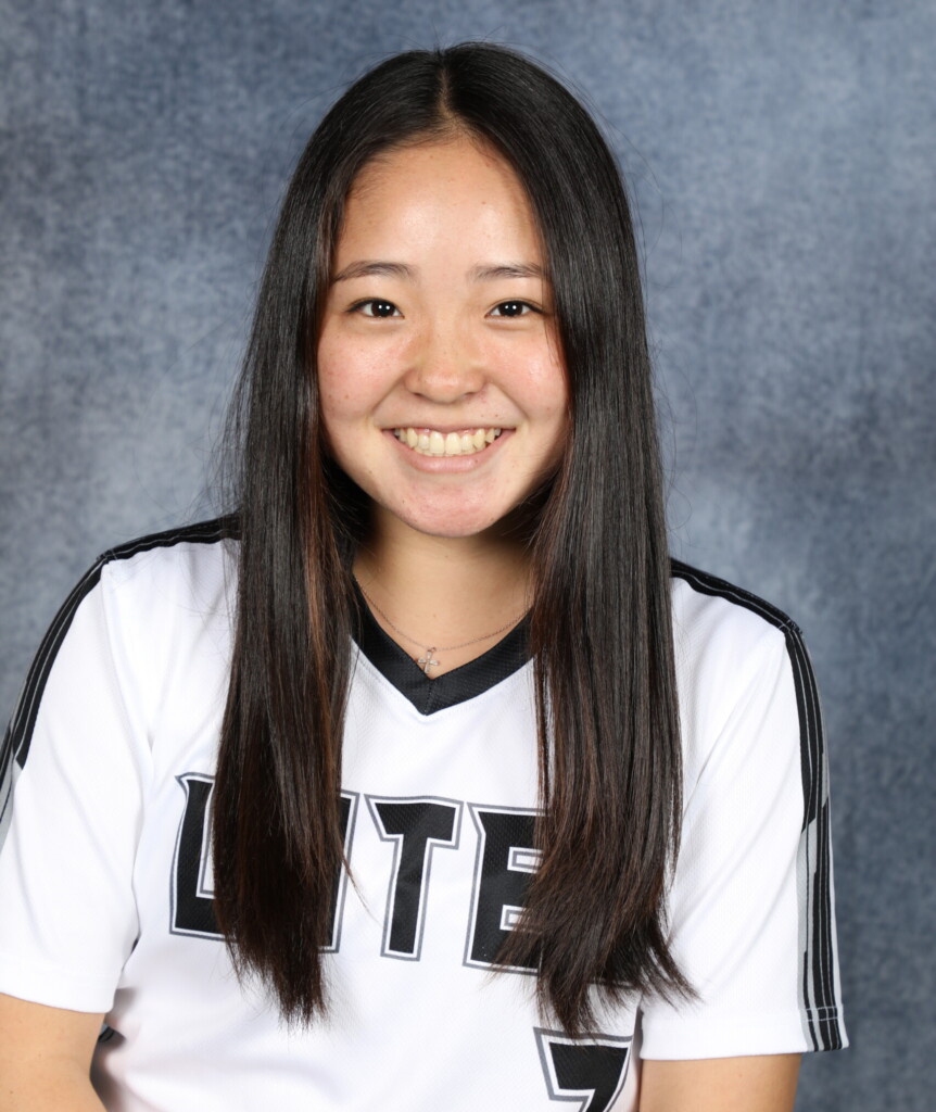 Rylie Wada smiles at the camera. Wada has long black hair and is wearing her black and white softball jersey.