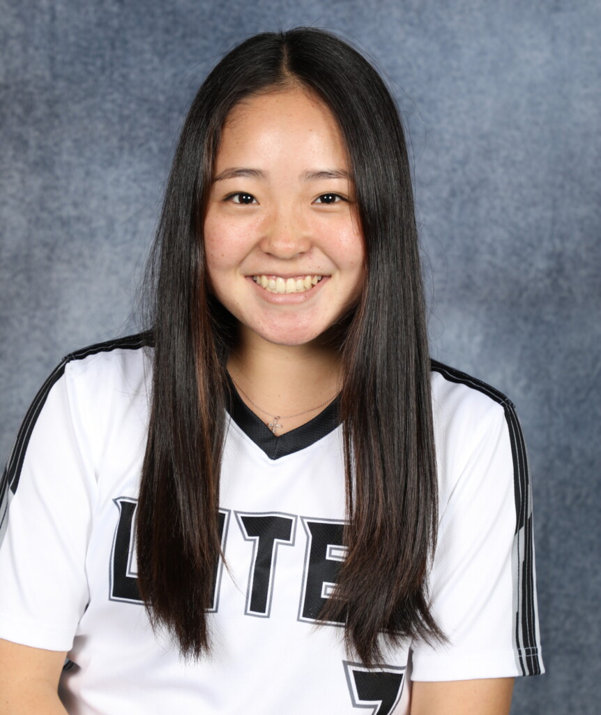 219A1621 Rylie Wada smiles at the camera. Wada has long black hair and is wearing her black and white softball jersey.