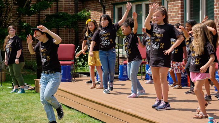 PLU student Ashton Allen leads campers in a dance number outside of Trinity Lutheran Church. The campers are on a stage, while Ashton performs in front of them from the grass.