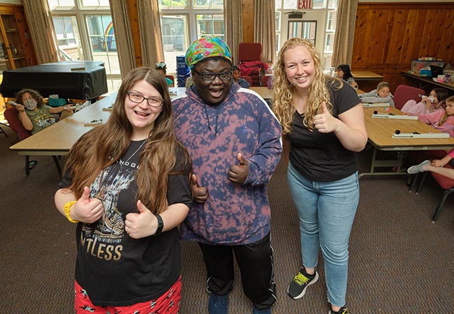 kaila-harris-zyreal-oliver-chandler-madison-ely Kaila Harris, Zyreal Oliver-Chandler and Madison Ely stand side by side giving a thumbs up to the camera. They are in a brown room, with students sitting at tables behind them.