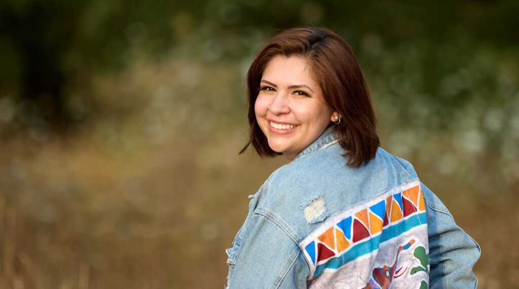Elizabeth Larios is pictured outside looking over her left shoulder. She is wearing a denim jacket with geometrical shapes on the back.