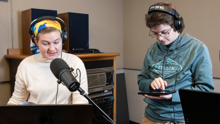 Z Rich sits in front of a microphone wearing a yellow bandana and cream colored sweater. Fulton Anderson-Bryant stands to her left touching an iPad. Both are wearing headphones.