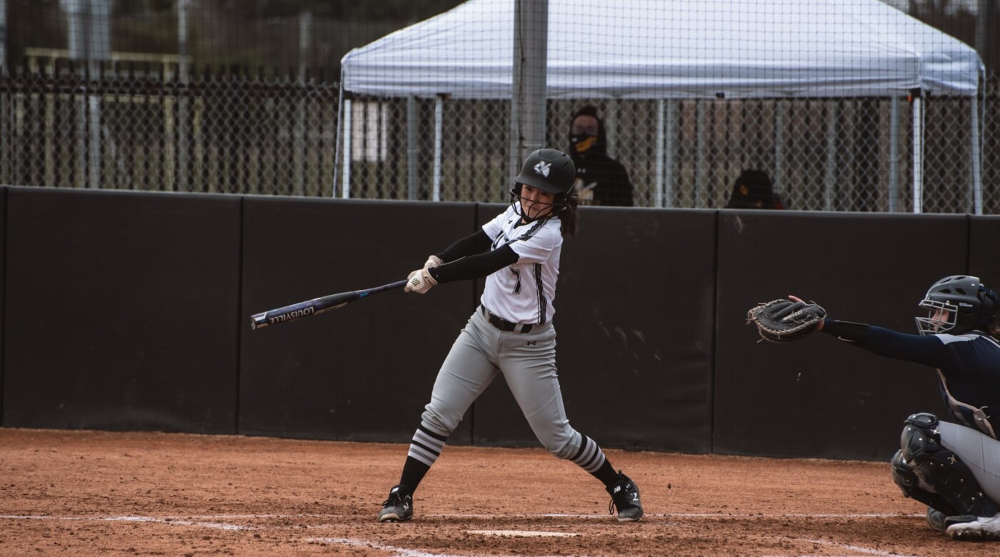 Rylie Wada is in her black and white softball jersey. She is swinging her bat with a catcher behind her.