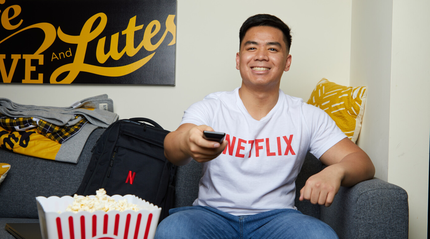 Adrian Ronquillo sits on a couch with a remote control in his hand. He's wearing a white shirt with the words 'Netflix' across the chest.