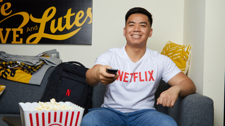 Adrian Ronquillo sits on a couch with a remote control in his hand. He's wearing a white shirt with the words 'Netflix' across the chest.