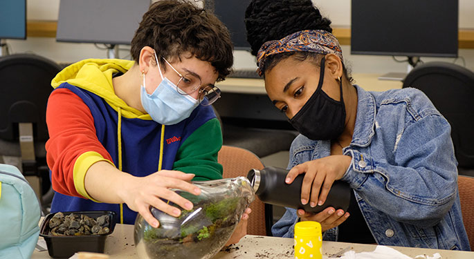 One students holds a glass jar filled with moss and sticks. Another student pours water into the jar.