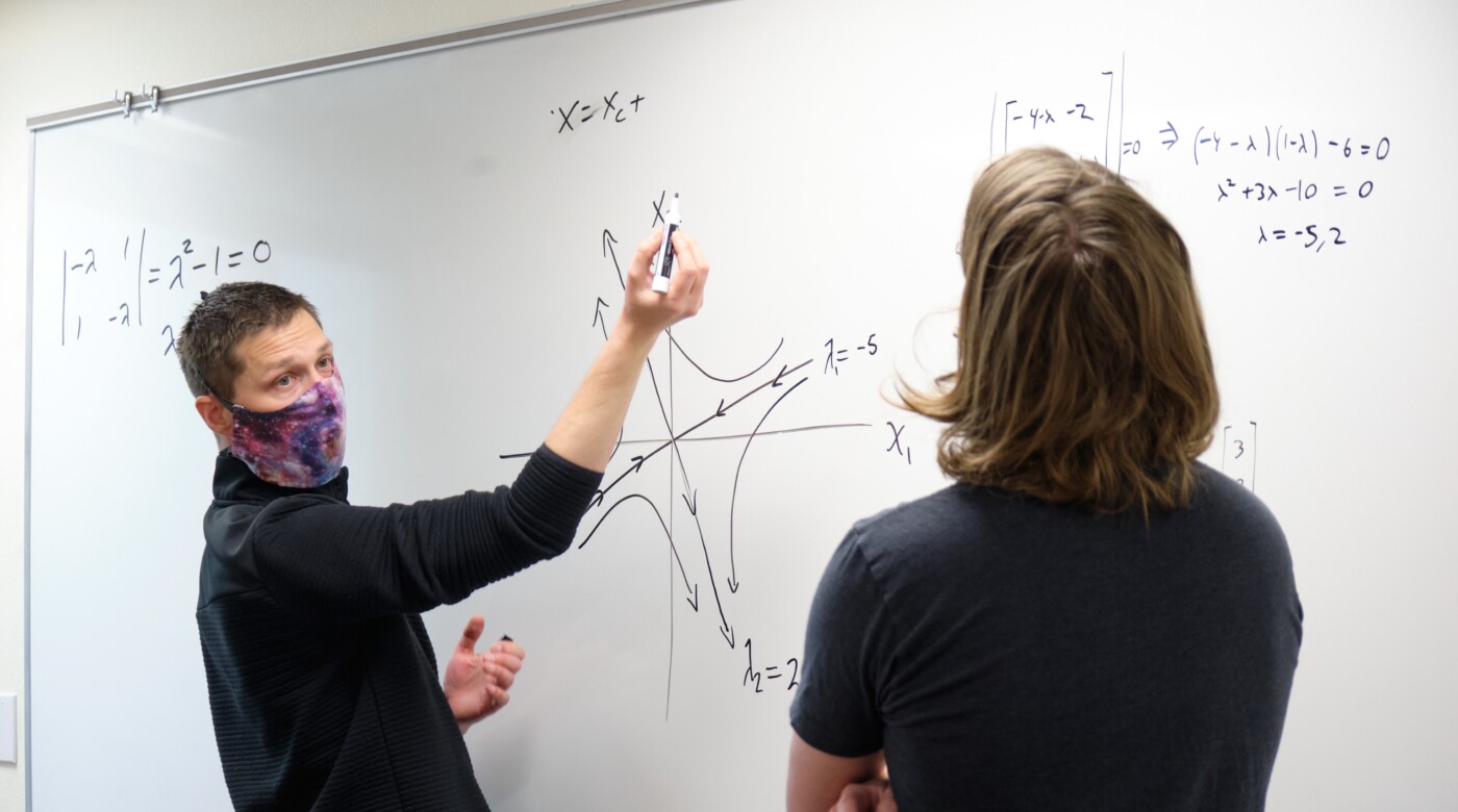 Professor Brent Underwood talks to a student with physics equations on a white board in front of them