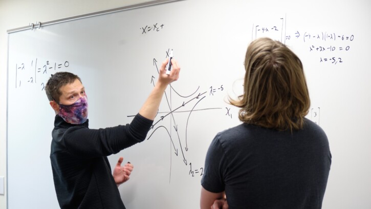 Professor Brent Underwood talks to a student with physics equations on a white board in front of them