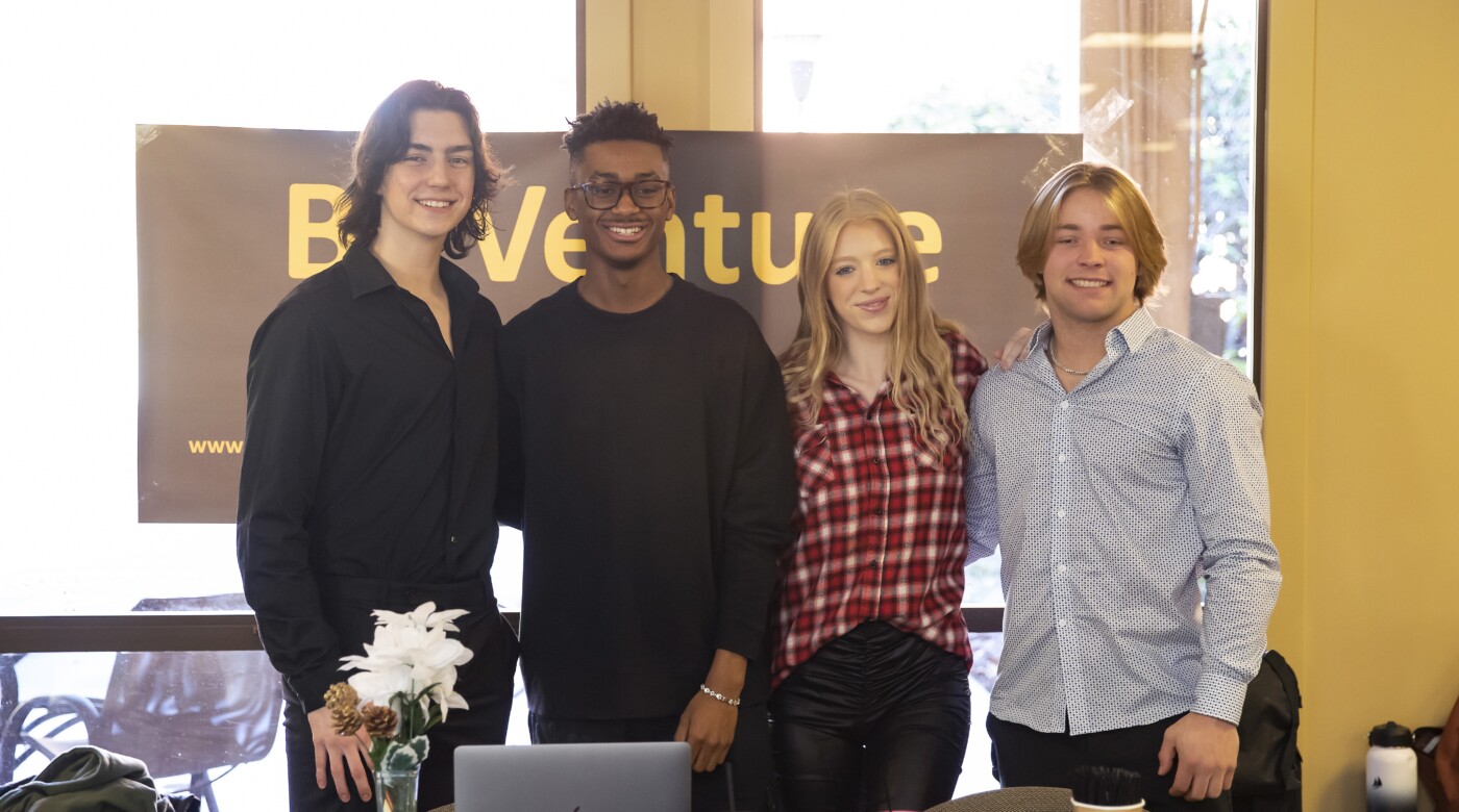 Four business students stand and smile together behind a table during PLU's Biz Venture Day