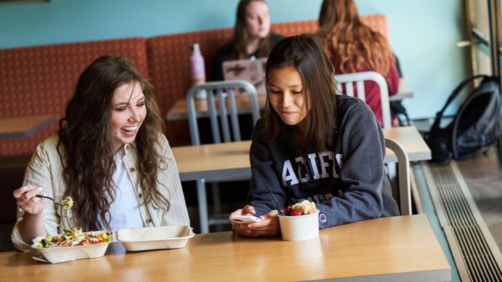 Two students sit at a table in PLU's Commons - one has a yogurt parfait and the other an entree with lots of vegetables.