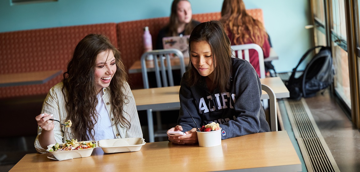 Two students sit at a table in PLU's Commons - one has a yogurt parfait and the other an entree with lots of vegetables.