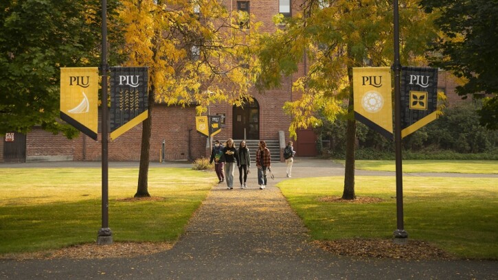 Four students walk down a sidewalk on campus, a brick building behind them. On either side of the sidewalk are black and yellow PLU banners hanging from the lamposts.