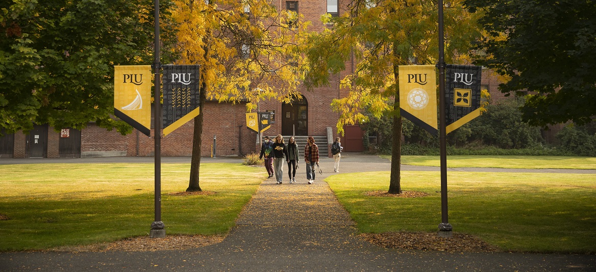 Four students walk down a sidewalk on campus, a brick building behind them. On either side of the sidewalk are black and yellow PLU banners hanging from the lamposts.