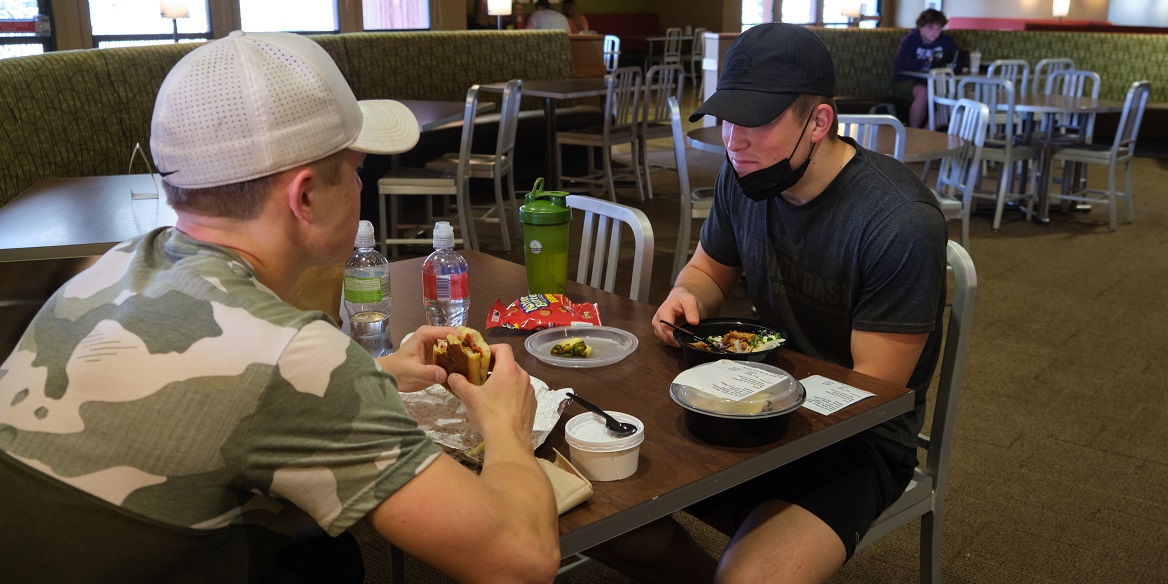 Two students, both wearing baseball caps, eat lunch in the PLU Commons