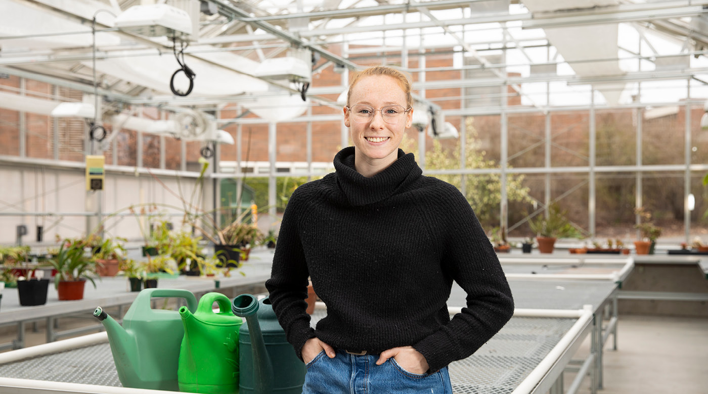 Sarah Davis, wearing a black sweater and blue jeans smiles for the camera while standing in PLU's greenhouse