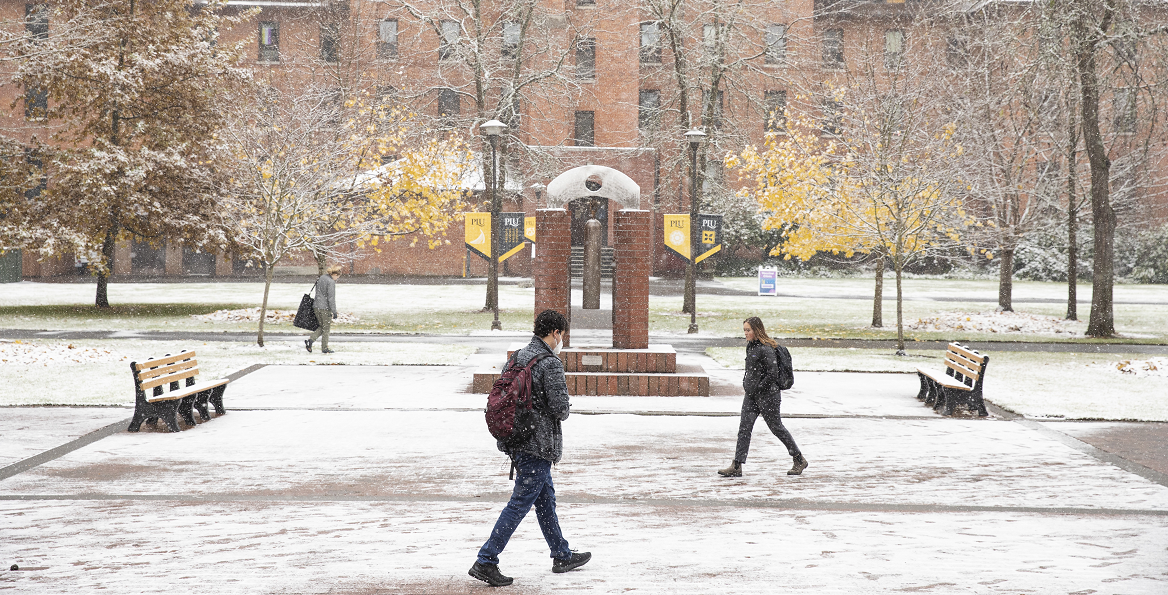 Students wearing backpacks walk across a snowy Red Square in the center of PLU's campus