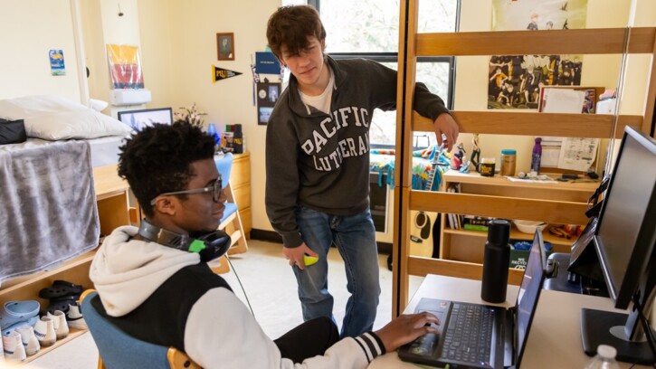 Two students in their residence hall room in Tingelstad. One is sitting at his desk under a lofted bed, with headphones around his neck. The other student stands near - both are looking at a laptop on the desk.