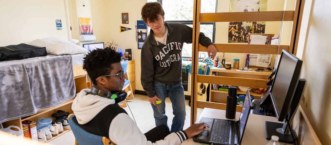 Two students in their residence hall room in Tingelstad. One is sitting at his desk under a lofted bed, with headphones around his neck. The other student stands near - both are looking at a laptop on the desk.