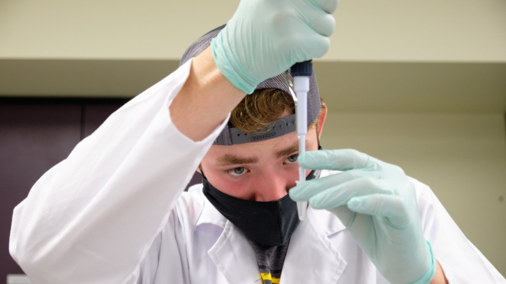 A student wearing a black mask, a blue baseball cap turned backward, and a white lab coat holds a pipette in front of their face as they practice adding liquid to a pipette.