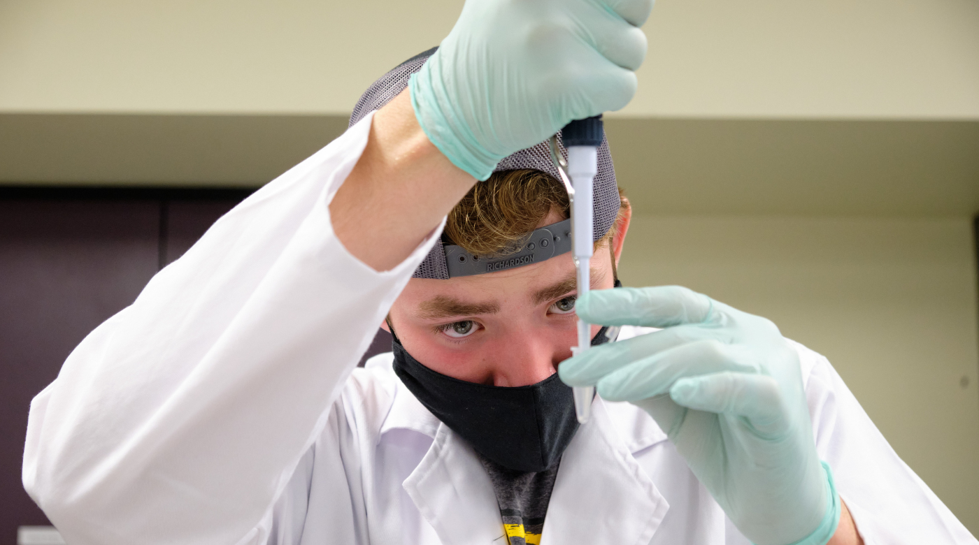 A student wearing a black mask, a blue baseball cap turned backward, and a white lab coat holds a pipette in front of their face as they practice adding liquid to a pipette.