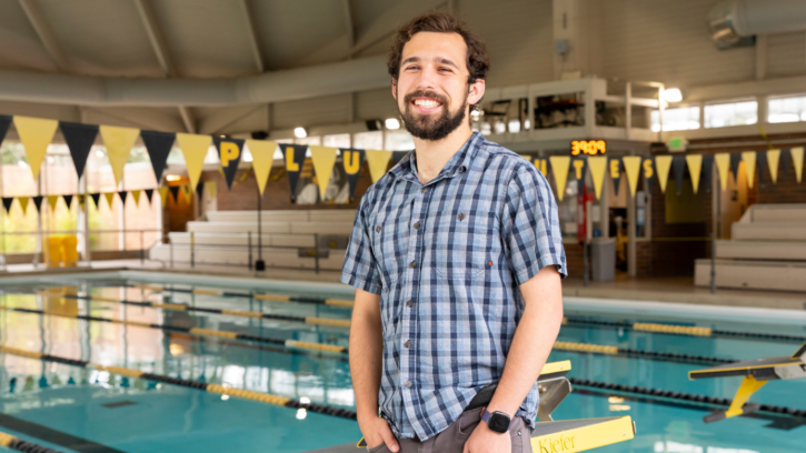 Student stands in front of the pool wearing a plaid shirt and smiles at the camera.