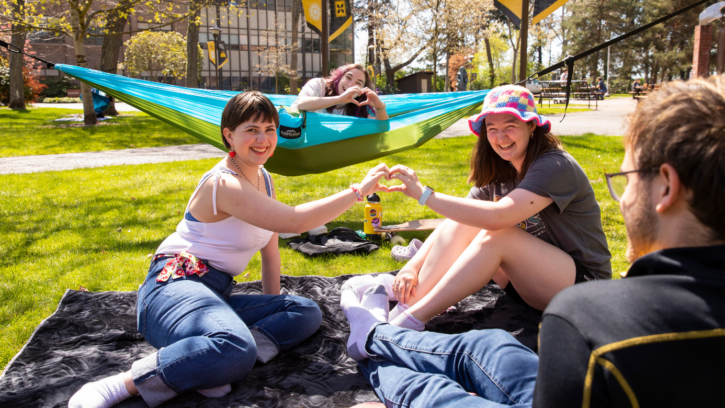 Four students are sitting outside on the grass. One student is sitting in a hammock and two students are making a heart with their hands.