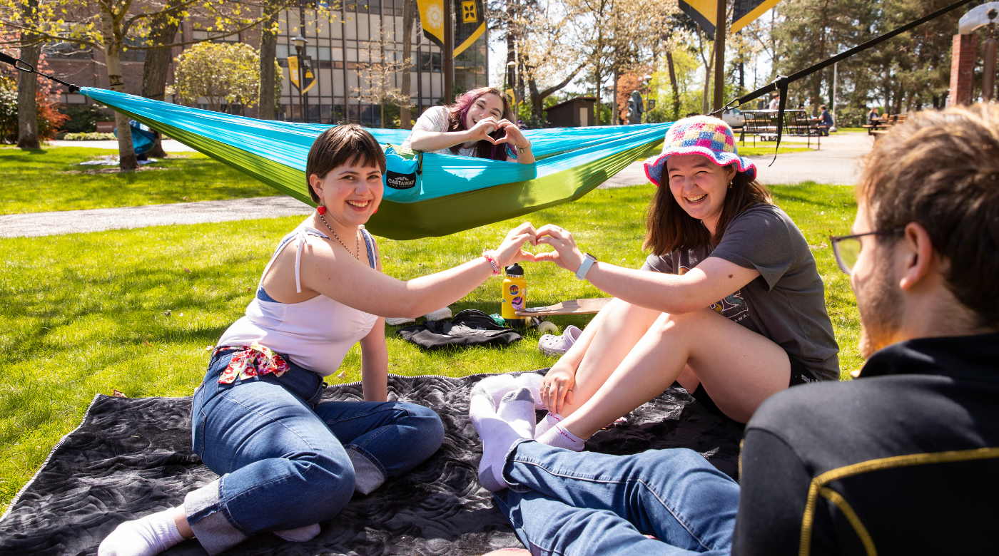 Four students are sitting outside on the grass. One student is sitting in a hammock and two students are making a heart with their hands.