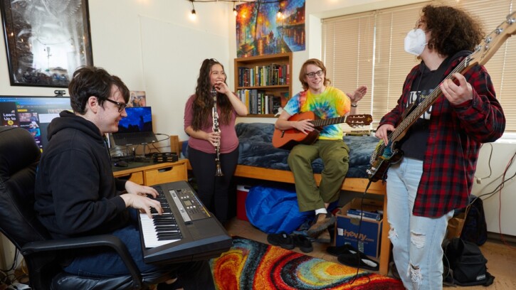 4 students with instruments (keyboard, guitar, clarinet, bass) hang out in a desk chair and on a bed in a PLU residence hall room.